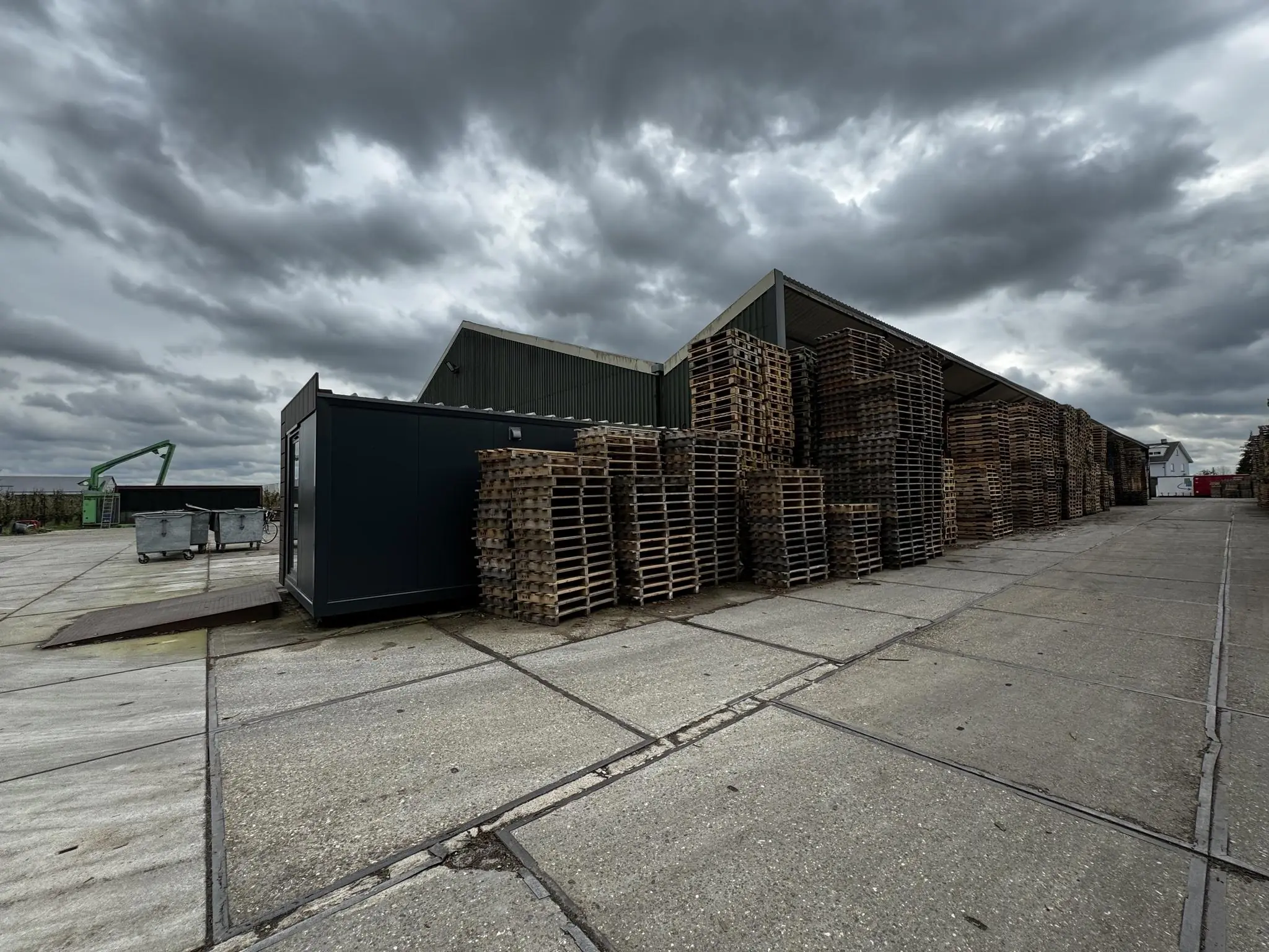 Opgeslagen houten pallets bij een loods aan de Graaf van Lynden van Sandenburgweg onder een dreigende, bewolkte lucht.